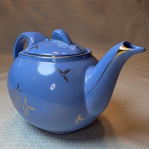May include: A light blue ceramic teapot with a lid, handle, and spout. The teapot is decorated with gold floral and leaf designs. The spout and rim have gold accents. The teapot is sitting on a light blue surface.