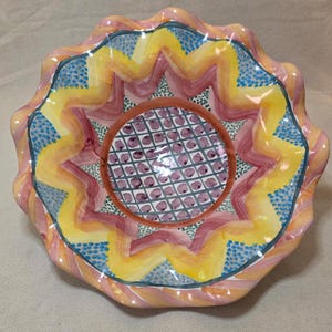 May include: A large, colorful ceramic bowl with a scalloped rim. The bowl is decorated with a geometric pattern of yellow, orange, pink, blue, and green. The center of the bowl features a grid pattern with purple dots.