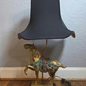 May include: A vintage table lamp with a gold-coloured horse statue base, featuring a green and brown saddle. The lamp has a black lampshade and a brass-coloured stem. The base is set on a dark red wooden platform.