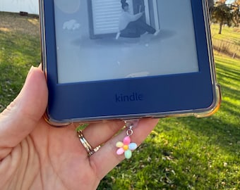 Puede incluir: Un lector de libros electrónicos Kindle azul con una funda transparente, que muestra un gráfico de una persona leyendo. Un amuleto de flor colorida cuelga del dispositivo. La palabra "Kindle" es visible.