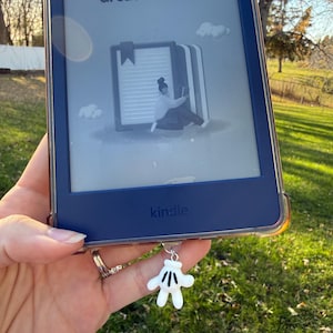 May include: A blue Kindle e-reader with a clear protective case, displaying the text "Editors' picks that are Great on Kindle." A white cartoon hand charm hangs from the device. The background is a grassy area with trees.