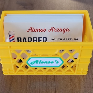 Könnte beinhalten: Eine gelbe Plastikkiste mit Visitenkarten. Die Karten zeigen "Alonso Arzaga" und "BARBER" mit einer Friseurstange. Auf der Kiste steht "Alonso's" auf der Vorderseite.