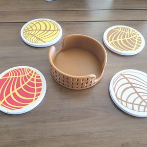 May include: A set of four round coasters with a white border and a brown, yellow, and red design, along with a matching brown coaster holder. The coasters feature a stylized pattern reminiscent of a concha pastry.