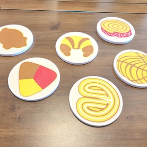 Op de afbeelding: Een verzameling witte, ronde onderzetters met kleurrijke illustraties van gebak. Ontwerpen zijn onder meer een croissant, een spiraalgebakje, een concha en andere gebakjes. De onderzetters zijn gerangschikt op een houten oppervlak.