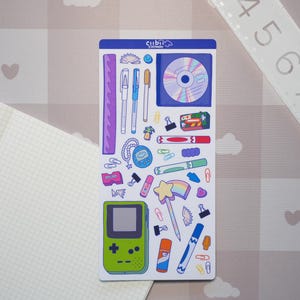 May include: A rectangular sticker sheet featuring various colorful illustrations including a green handheld game console, a CD, a star wand, pens, paper clips, and a ruler. The sheet measures approximately 8 inches in length as shown by the ruler.