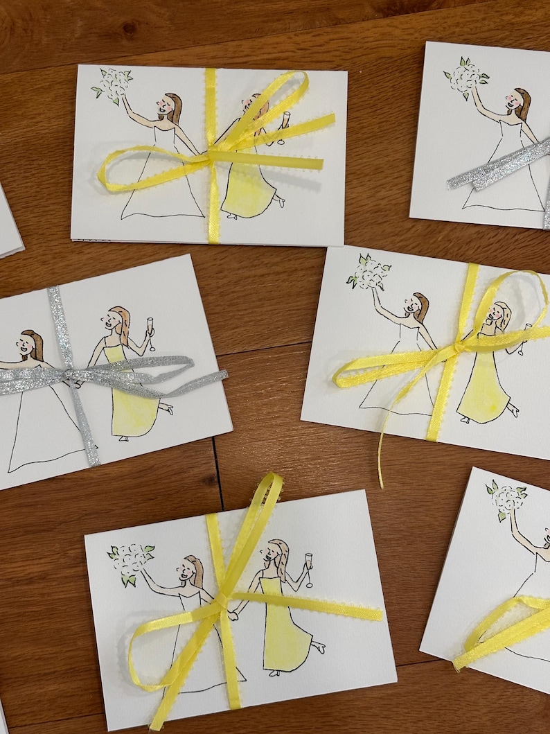 Puede incluir: Una colecci&oacute;n de tarjetas rectangulares blancas con un dibujo lineal de dos figuras con vestidos, una sosteniendo un ramo y la otra una copa. Algunas tarjetas est&aacute;n atadas con cinta amarilla brillante, otras con cinta plateada.
