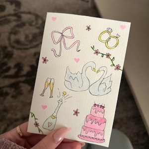 Puede incluir: Una tarjeta rectangular blanca con ilustraciones dibujadas a mano en tinta y acuarela. El diseño incluye un lazo rosa, dos anillos dorados, un par de cisnes blancos, dos copas de champán, una botella, un pastel rosa de varios pisos y corazones.