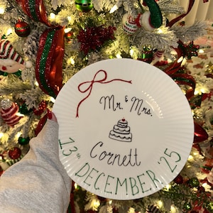 Puede incluir: Un plato blanco con un lazo rojo, las palabras "Mr. & Mrs." y una ilustración de pastel. El plato también incluye los nombres "Cornett" y "Diciembre 25" en verde, con la fecha "13th" en verde.
