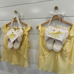 Puede incluir: Dos conjuntos de tops sin mangas de color amarillo claro con hombros con volantes, cada uno combinado con un par de pantuflas de piel sint&eacute;tica color crema. Cada conjunto cuelga de una percha de madera con una cinta blanca y una tarjeta.
