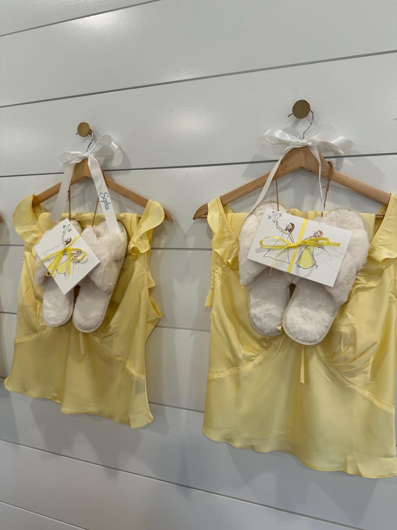 Puede incluir: Dos conjuntos de tops sin mangas de color amarillo claro con hombros con volantes, cada uno combinado con un par de pantuflas de piel sint&eacute;tica color crema. Cada conjunto cuelga de una percha de madera con una cinta blanca y una tarjeta.
