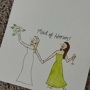 Puede incluir: Una tarjeta de felicitaci&oacute;n blanca con una ilustraci&oacute;n dibujada a mano de dos figuras tomadas de la mano. Una figura lleva un vestido blanco y sostiene un ramo, mientras que la otra lleva un vestido verde y sostiene una copa. El texto Maid of Honor? aparece arriba.
