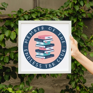 Könnte beinhalten: Weiß gerahmter Druck mit einem kreisförmigen Design mit einem Stapel von Büchern in Rosa, Türkis und Marineblau. Der Text lautet "MEMBER OF THE ENDLESS TBR CLUB" in Marineblau um den rosa Kreis. Der Hintergrund ist eine grüne Pflanze.