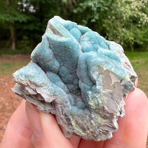 May include: A close-up of a teal-colored mineral specimen. The mineral has a layered, textured appearance with a mix of teal and gray hues. The specimen is held in a hand, with a blurred green background of trees and grass.