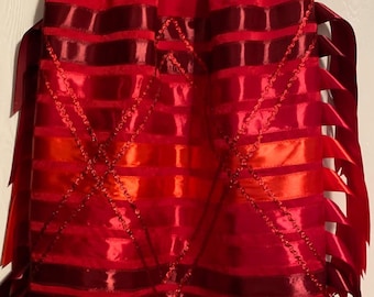 May include: A vibrant red skirt with horizontal bands of varying shades of red and orange. The skirt is adorned with decorative ribbon and chain accents, and has long, pointed ribbon details on the sides.