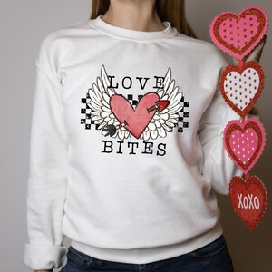 May include: White sweatshirt with a red heart with wings and an arrow through it. The text "LOVE BITES" is printed below the heart. The person is holding a string of pink and red glitter hearts with white polka dots. The last heart has the text "XOXO" printed on it.