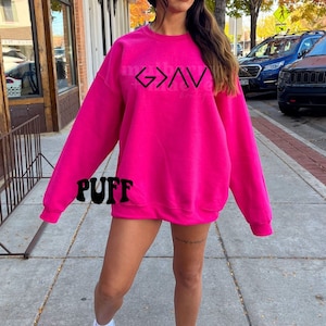 May include: A pink sweatshirt with the word "G>A" printed on the front. The sweatshirt has a crew neck and long sleeves. The word "PUFF" is printed on the sleeve.