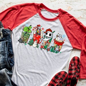 May include: A raglan-style shirt with a gray body and red sleeves. The shirt features a colorful graphic of Nightmare Before Christmas characters in coffee cup designs. A pair of blue jeans and red and black plaid shoes are next to the shirt.