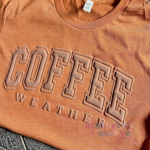 May include: A heathered orange t-shirt featuring the word COFFEE in large, raised, light-colored block letters with a secondary line of smaller, raised text reading WEATHER below it. A white size tag is visible at the inner neckline.