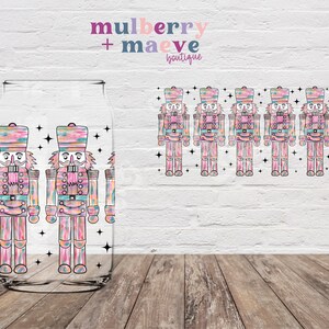 May include: A clear glass tumbler with a colorful, abstract design of a nutcracker. The design features pink, blue, and green hues with black outlines. The tumbler is surrounded by a white brick wall and a wooden floor.