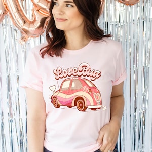 May include: A pink t-shirt with a retro-style illustration of a pink and white Volkswagen Beetle with the text "Love Bug" in a groovy font.