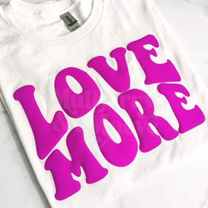 May include: A white short-sleeved t-shirt featuring the text LOVE MORE printed in a large, bold, bright pink font across the chest. The lettering has a rounded, wavy style. The garment includes a standard crew neckline and a visible brand label.