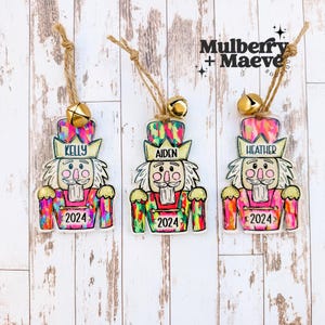 May include: Three colorful nutcracker ornaments with gold bells and the names Kelly, Aiden, and Heather. Each ornament has the year 2024 printed on it.