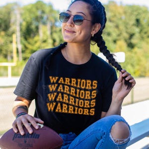May include: A black short-sleeve t-shirt featuring the word WARRIORS printed four times in a stacked, orange-outlined font. The shirt is worn with blue jeans and paired with a brown leather football held in the hand.