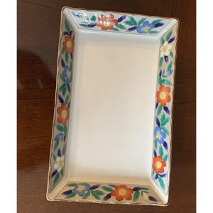 May include: Rectangular ceramic serving tray with a cream-colored interior and a floral border. The border features orange, blue, and white flowers with green leaves on a light beige background. The tray has a gold trim.