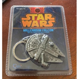 Op de afbeelding: Een zilverkleurige metalen replica sleutelhanger van de Millennium Falcon uit Star Wars. De sleutelhanger zit in een transparante plastic verpakking met het Star Wars logo en de tekst "Millennium Falcon". De verpakking vermeldt ook "For ages 14+".