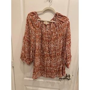 May include: A long-sleeved blouse with a rust-colored and white pattern. The blouse has a V-neckline and buttons down the front. The sleeves are three-quarter length. The blouse is hanging on a white hanger.