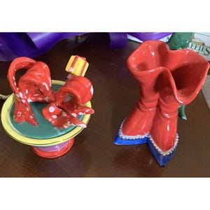 May include: Two ceramic decorative pieces. One is a red bow with white polka dots on a pedestal. The other is a pair of red boots with blue and gold accents. Both pieces are brightly colored.