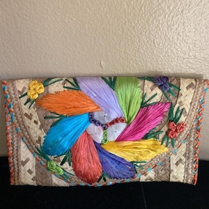 Puede incluir: Bolso de mano tejido con un diseño floral colorido. La flor central tiene pétalos en tonos azules, naranjas, morados, verdes, rosas y amarillos. Los bordes están adornados con costuras naranjas y azules. Hecho de paja trenzada.