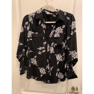Pode incluir: Camisa preta com estampa floral branca. A camisa de manga comprida tem dois bolsos frontais e mangas enroladas. A camisa está pendurada em um cabide bege.