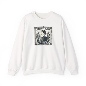 Puede incluir: Sudadera blanca con un estampado gráfico de una mujer leyendo un libro. El gráfico está en un marco negro con el texto "Sapphic Book Club".