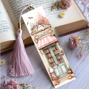 May include: A decorative bookmark featuring a bookstore illustration with a pink awning and teal doors. The bookmark has a tassel and is decorated with bats, pumpkins, and a ghost. The word "BOOKSTORE" is printed on the awning.