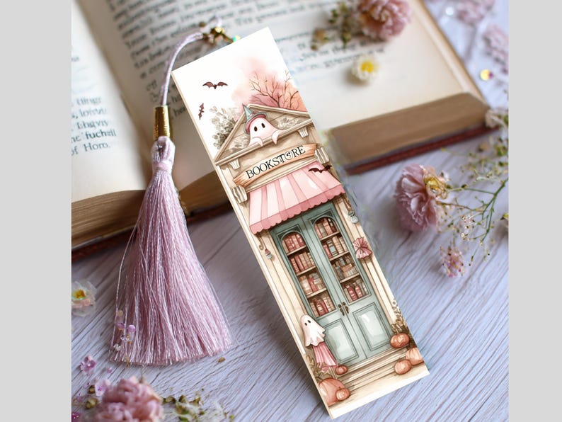 May include: A decorative bookmark featuring a bookstore illustration with a pink awning and teal doors. The bookmark has a tassel and is decorated with bats, pumpkins, and a ghost. The word "BOOKSTORE" is printed on the awning.