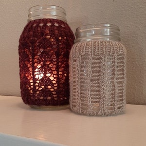 Pode incluir: Dois frascos de vidro com capas de malha. O frasco à esquerda tem uma capa de malha bordô com um padrão de renda. O frasco à direita tem uma capa de malha bege com um padrão de nervuras.
