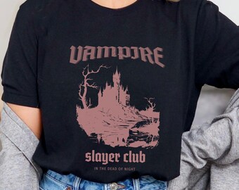 May include: Black t-shirt with a pink graphic of a castle and the words "VAMPIRE slayer club" in a gothic font. The phrase "IN THE DEAD OF NIGHT" is below the club name. The shirt is made of a soft material.