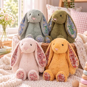 May include: Four plush stuffed bunnies in sage green, olive green, soft pink, and golden yellow. Each bunny features long ears and foot pads with a floral pattern. The names Riley, Julia, Olivia, and Emily are embroidered on the inner ears.
