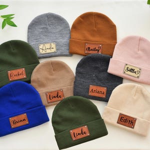 May include: A collection of personalized beanies in various colors, including gray, brown, pink, and blue. Each beanie features a rectangular leather patch with a name in script lettering. The beanies are arranged on a white surface.