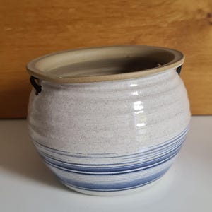 May include: A ceramic pot with a rounded shape, featuring a speckled white exterior with horizontal blue stripes. The rim and interior are a light tan colour. Small black handles are attached to the sides.
