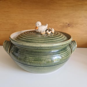 May include: Green ceramic casserole dish with a lid, featuring a duck and ducklings figurine on top. The dish has a ribbed texture and two handles. The lid and dish are green with a cream-coloured rim.