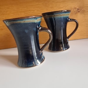 May include: Two ceramic mugs with a dark blue glaze and brown handles. The mugs have a unique, curved shape and a contrasting brown rim. The background is a light wood surface.