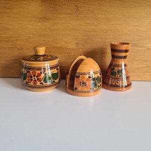 May include: A three-piece wooden set comprising a sugar bowl, napkin holder, and vase. Each item features orange, green, and brown floral patterns. The set is displayed on a white surface with a wooden backdrop.