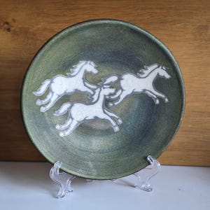 May include: A circular, green ceramic plate featuring three white horse figures in motion. The plate has a dark green rim and is displayed on a clear plastic stand. The horses are depicted with detailed manes and tails.