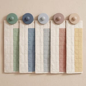 May include: A set of five quilted fabric wall organizers with wooden knobs. Each organizer has a different colored stripe: green, blue, gray, pink, and yellow. The background is a neutral beige.
