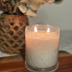 May include: A burning candle in a clear glass jar with a diamond pattern. The candle has two wicks and a warm glow. The candle sits on a wooden surface. A woven vase with dried flowers is in the background.