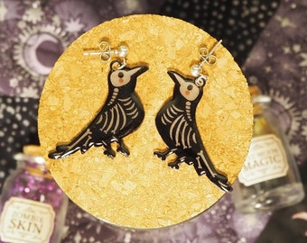 May include: A pair of black skeleton bird earrings with silver hooks. The earrings are on a cork background.