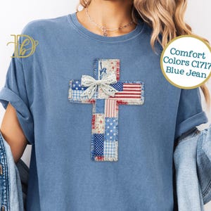 May include: Blue jean Comfort Colors t-shirt featuring a patchwork cross design with patriotic patterns and a white lace bow. The shirt has rolled-up sleeves and a relaxed fit, perfect for casual wear.