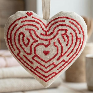 May include: A heart-shaped, off-white fabric ornament with a red labyrinth design. The ornament has a small loop for hanging and is displayed in front of a wooden background. A stack of folded fabric and a mug are in the background.